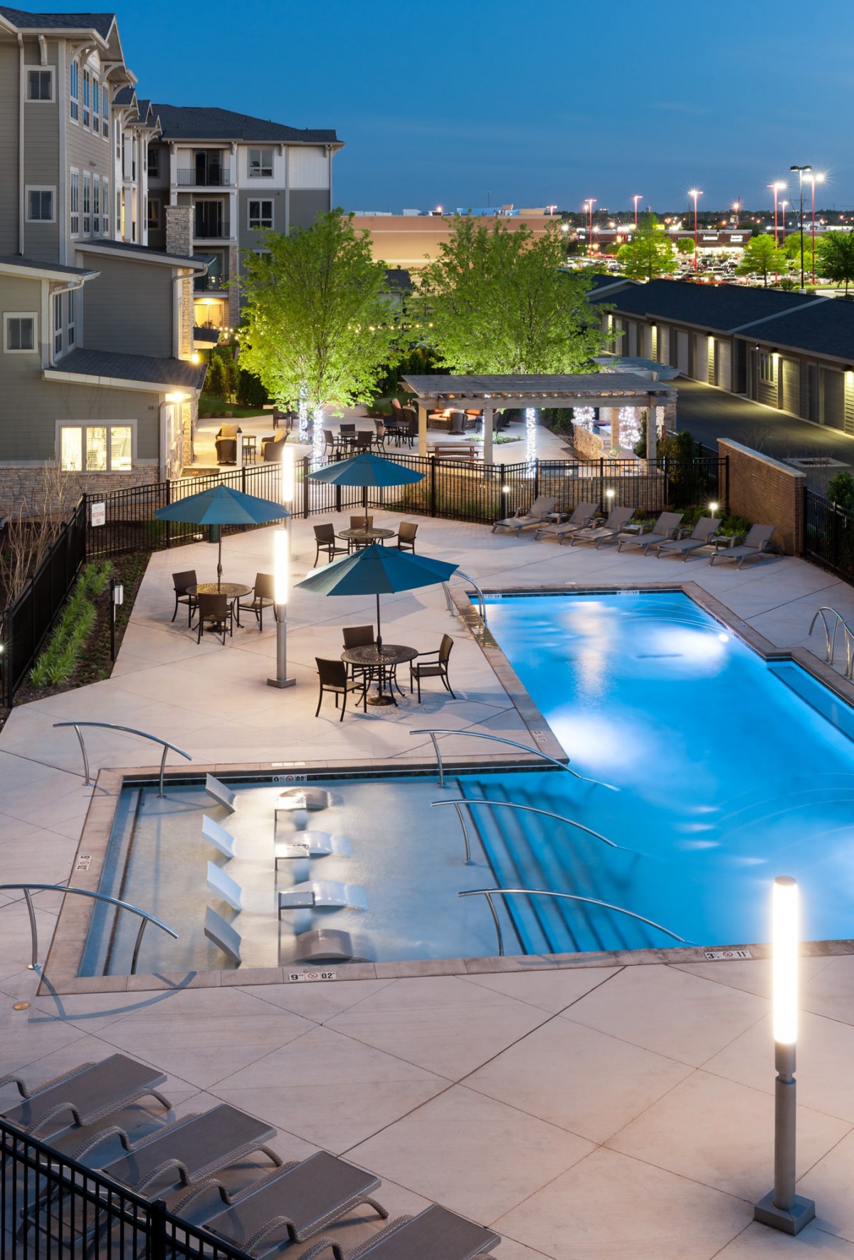 An inviting outdoor swimming pool area in a residential complex, featuring patio furniture and umbrellas under garden lights during the evening.