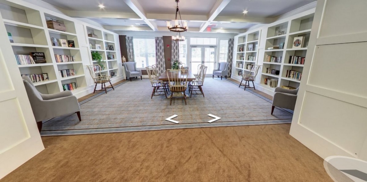 A spacious and elegant room with bookshelves, a dining table, and chairs, featuring large windows and soft lighting.