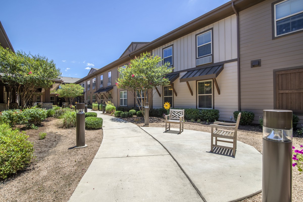 A sunny pathway leading to a building with outdoor seating and landscaping.