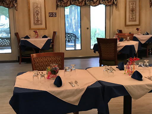 A cozy restaurant interior featuring elegantly set tables with blue and white tablecloths, decorated with flowers and a view of the outdoors.