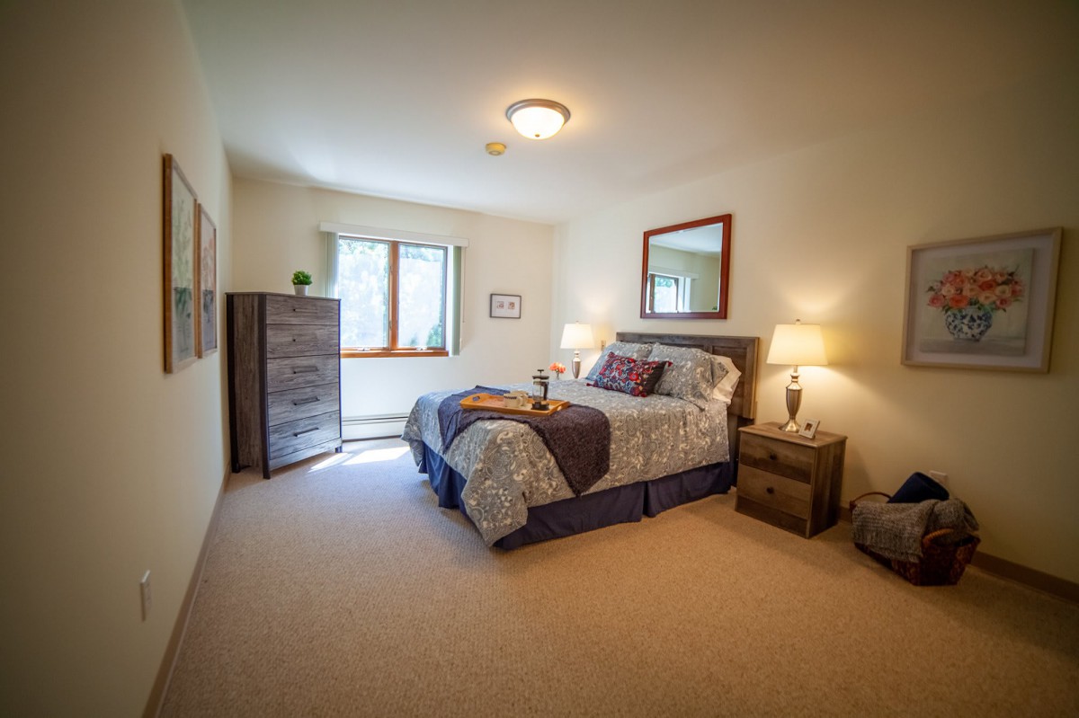 A cozy bedroom with a neatly made bed, two bedside lamps, and a dresser by the window.