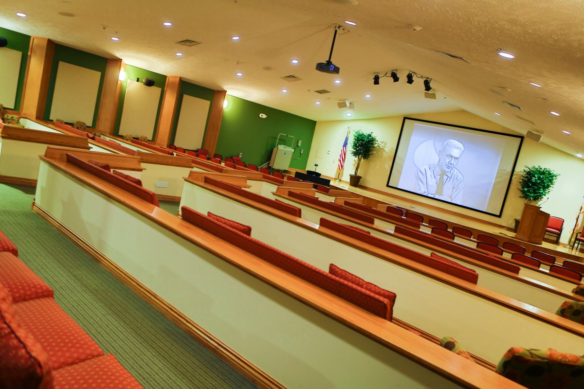 An empty lecture hall with rows of desks facing a large projector screen.