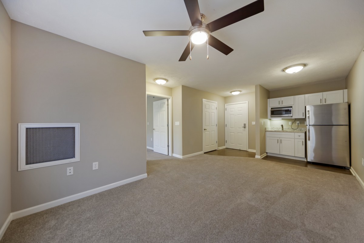 Interior view of a carpeted living room with a ceiling fan, leading into a small kitchen area.