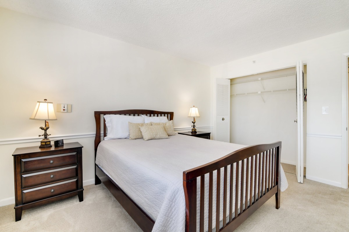 A minimalist bedroom with a wooden bed frame and white bedding, featuring a nightstand and lamp.
