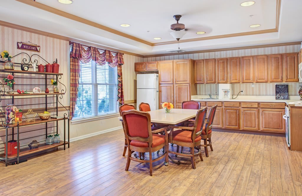 A spacious kitchen and dining area featuring wood cabinets, a large table with red upholstered chairs, and a window with plaid curtains.