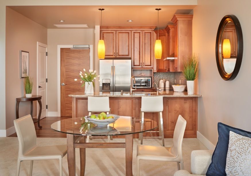 A modern kitchen and dining area featuring wood cabinets, stainless steel appliances, and a glass table set against a backdrop of neutral tones.
