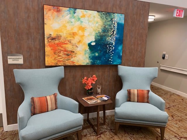 A cozy lounge area with two blue armchairs, a small table with magazines and flowers, and a colorful abstract painting on the wall.