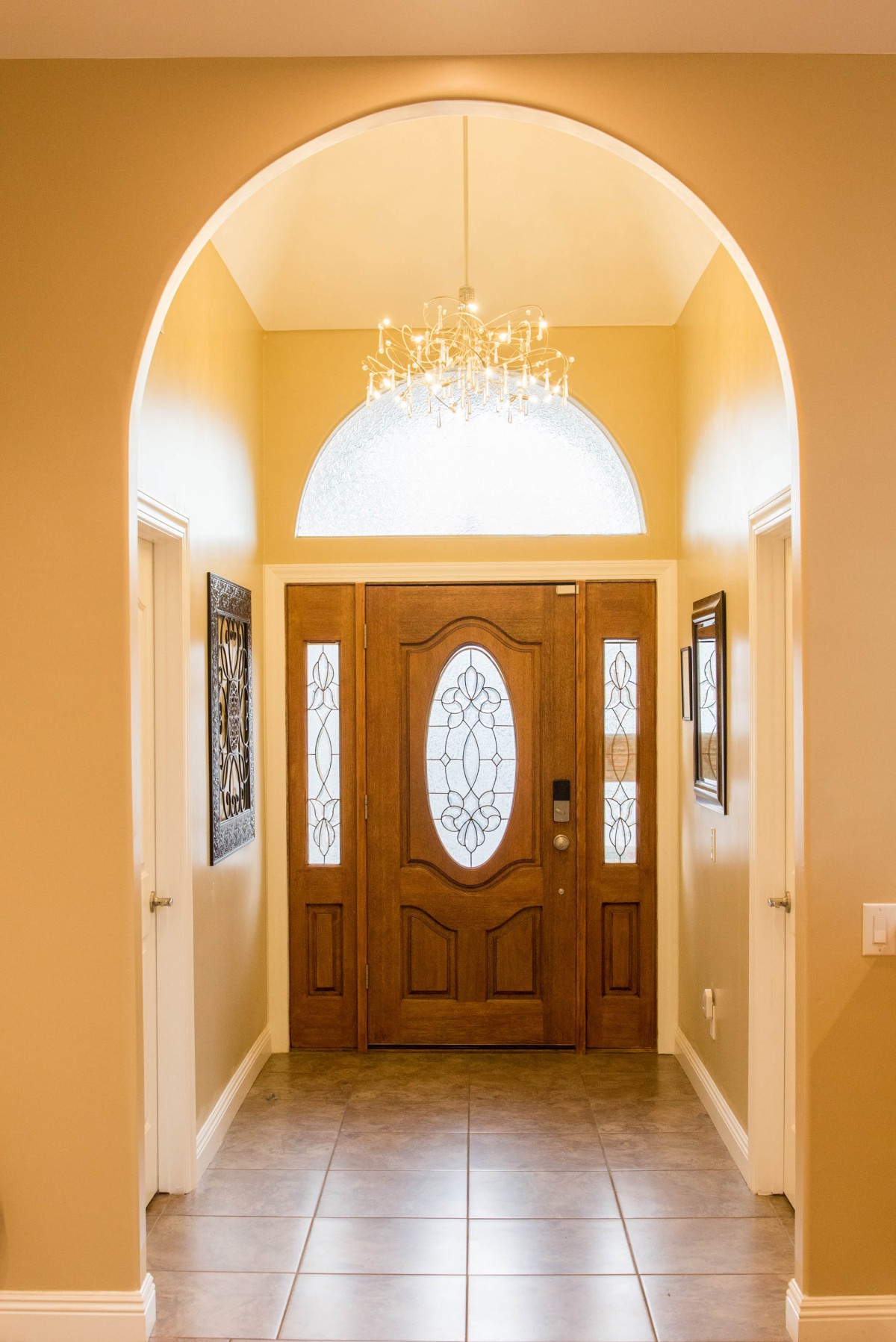 A warmly lit entryway featuring a wooden front door with decorative glass panels and an elegant chandelier.