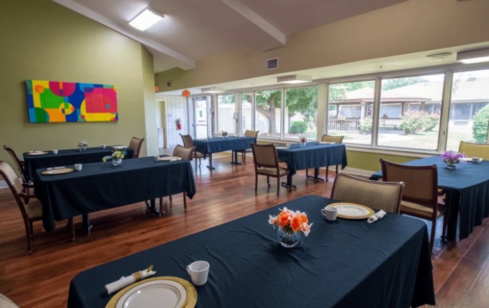 A spacious dining room with neatly arranged tables and chairs, featuring large windows and colorful artwork on the wall.