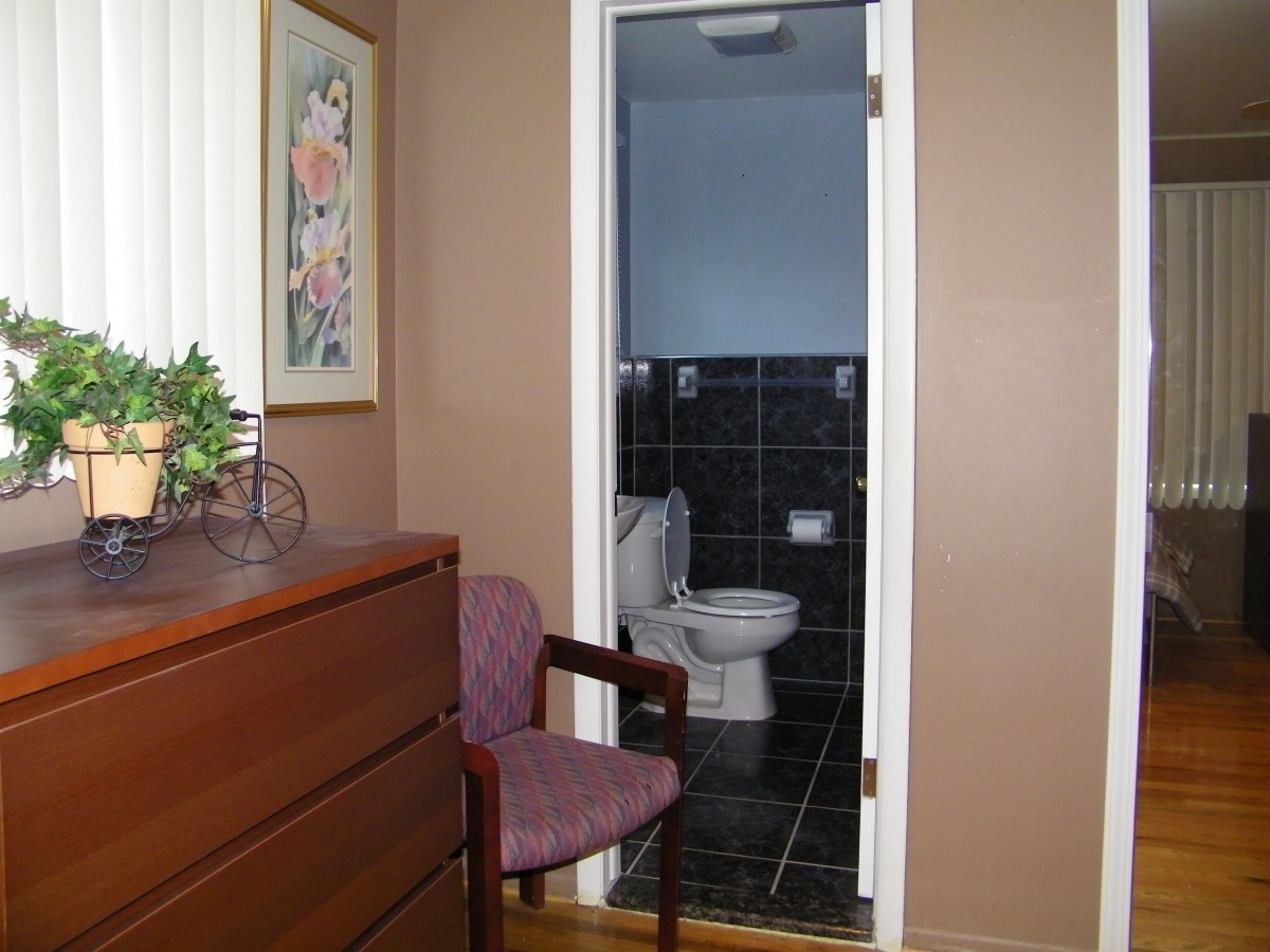 A bedroom with a wooden dresser and chair, leading to a small bathroom.