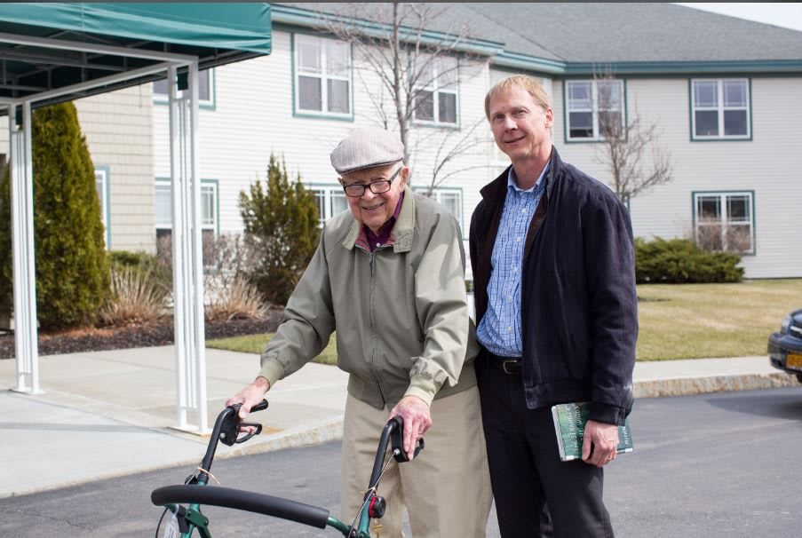 An elderly man with a walker is accompanied by a caregiver outdoors.
