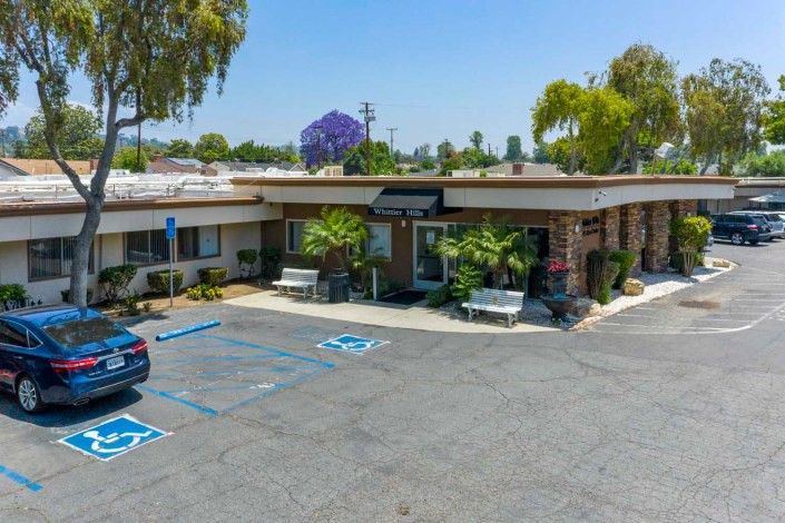 A small commercial building with a parking lot and greenery on a sunny day.