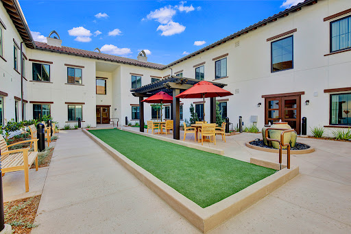 A serene outdoor courtyard featuring a modern design, with a green lawn and shaded dining areas under red umbrellas.