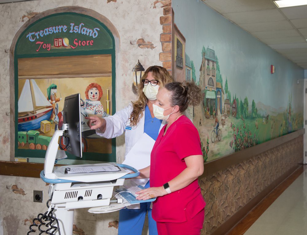 Two nurses collaborate over medical records in a children's hospital corridor decorated with vibrant murals.