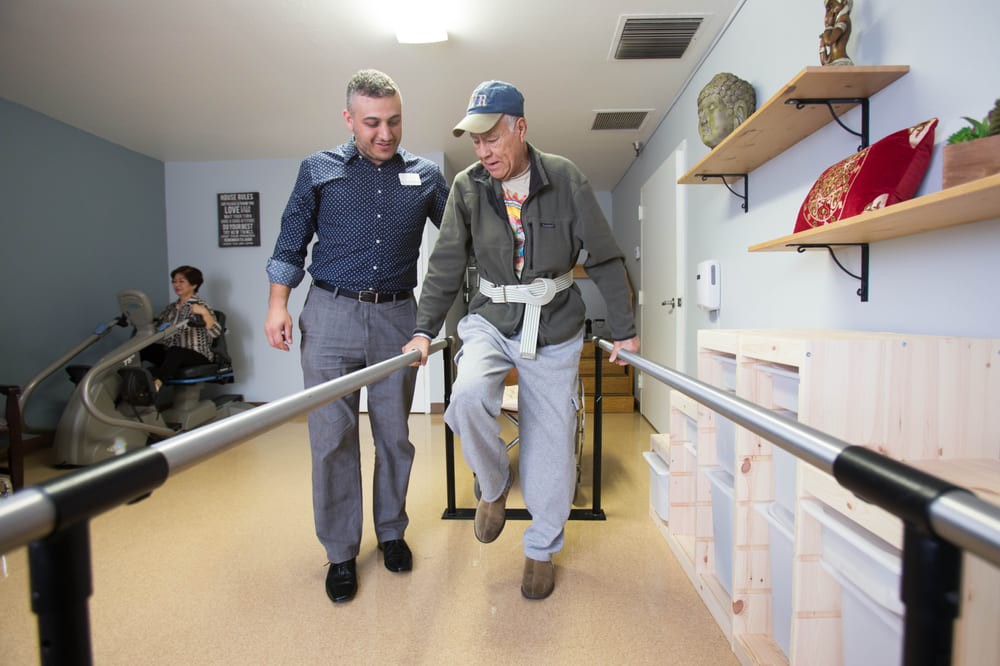 An elderly man receives support during a physical therapy session.