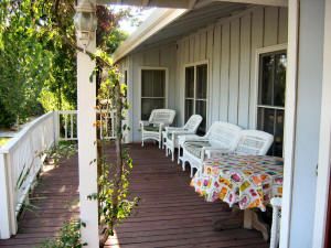 A sunny porch with white chairs, a table covered with a floral cloth, and plants climbing around the railing.