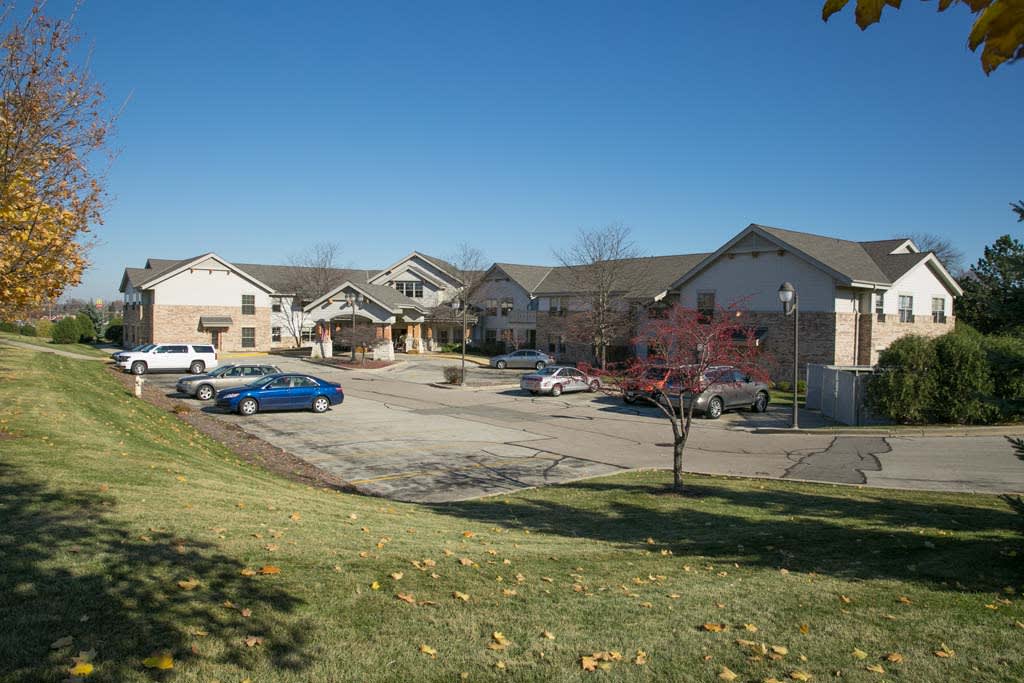 A view of a building complex with a parking lot and cars, surrounded by grass and trees under a clear blue sky.