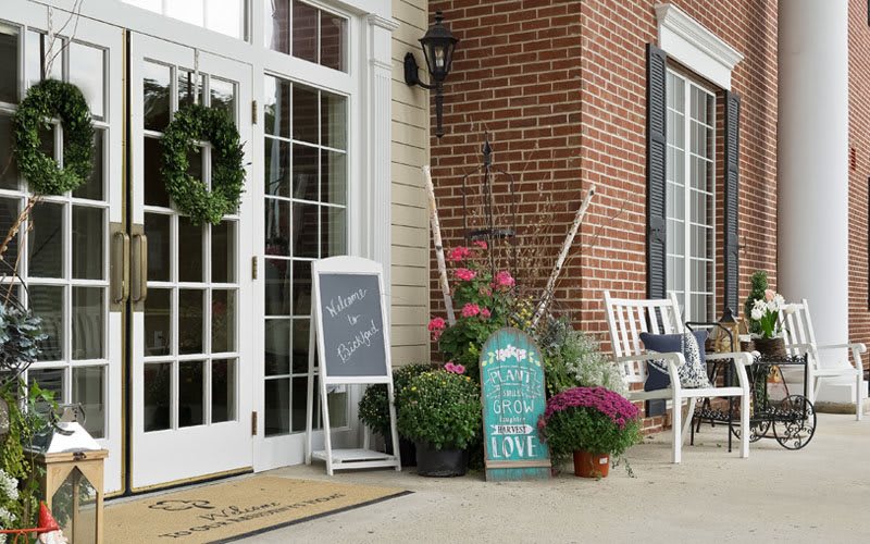 A charming storefront with a chalkboard sign, surrounded by flowers and garden tools, in front of a brick building.
