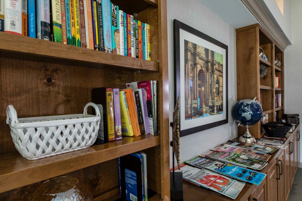 A cozy reading nook with a wooden bookshelf filled with colorful books, a decorative basket, and framed artwork on the wall. Nearby, magazines are neatly arranged next to a small globe.