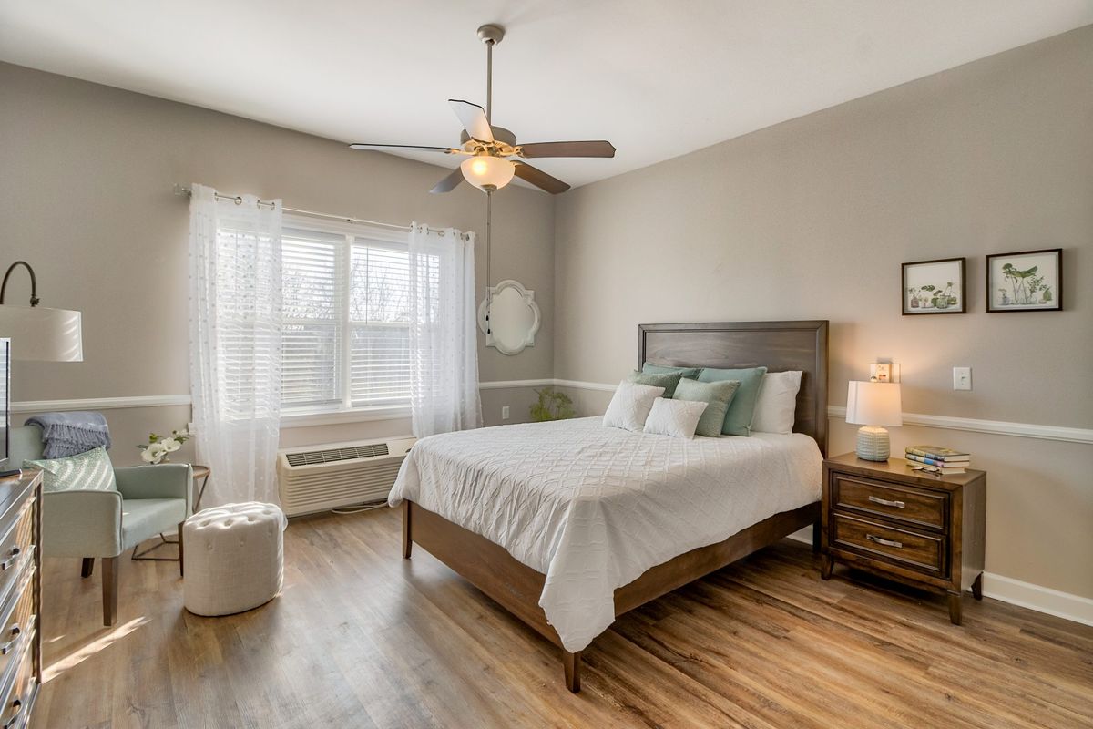 A modern bedroom featuring a bed with pillows, a nightstand with a lamp, and a ceiling fan. The room has hardwood floors and large windows with sheer curtains.