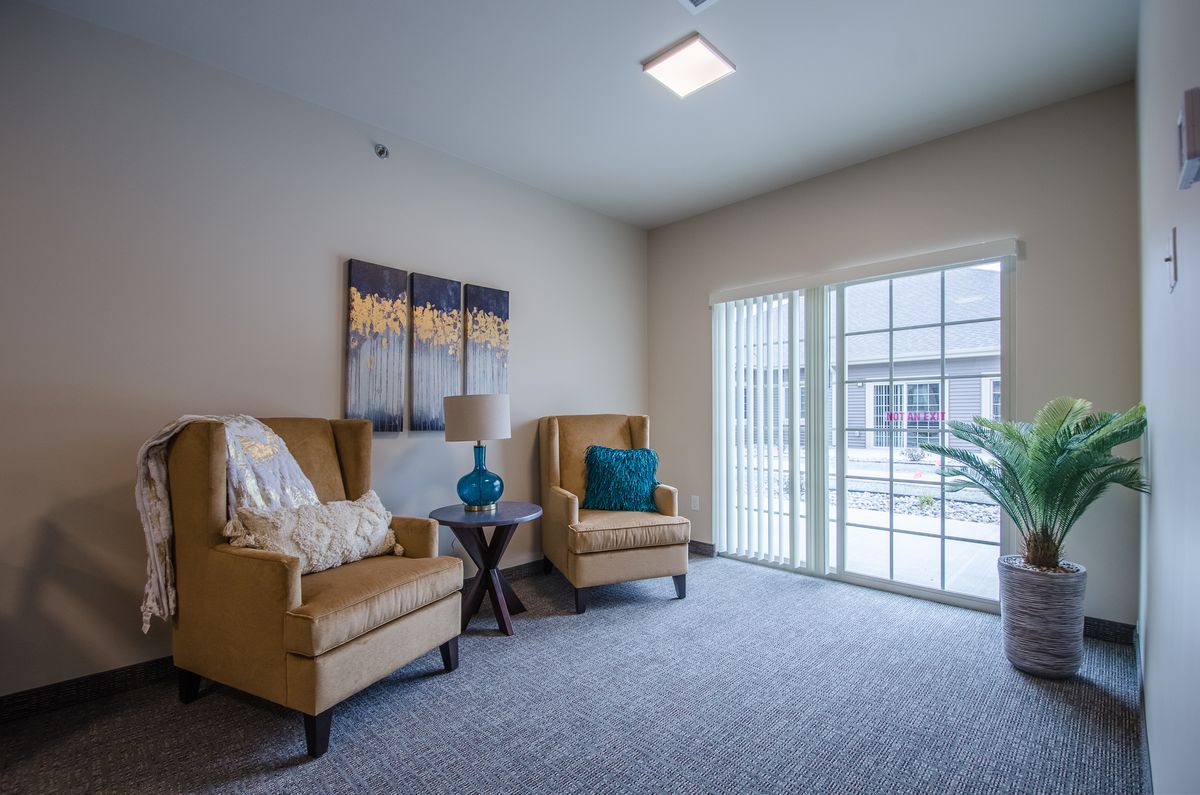A cozy living room with two armchairs, a side table with a lamp, artwork on the wall, and plenty of natural light streaming in through large windows.