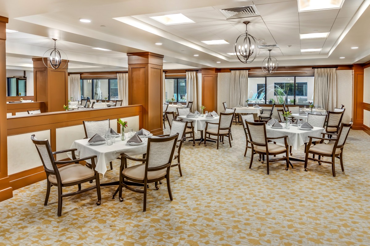 An elegant restaurant dining room with neatly arranged tables and chairs, featuring a chandelier and carpeted flooring.
