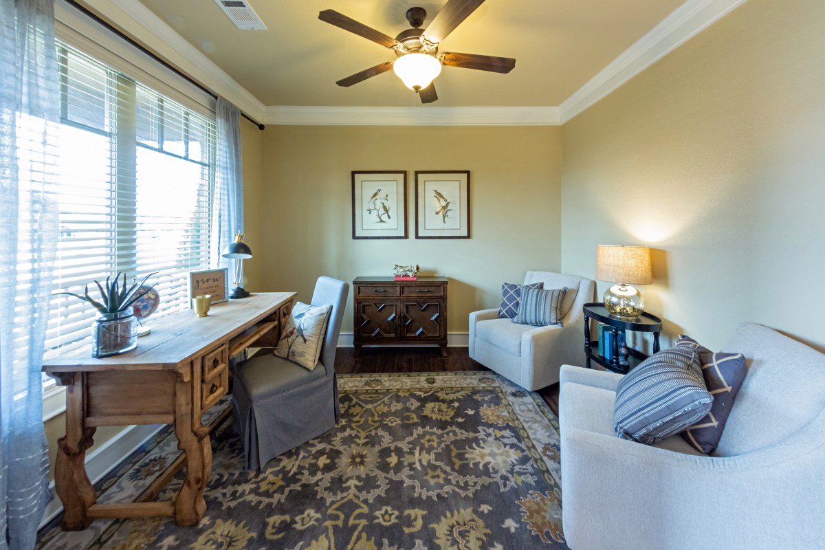 A cozy living room with elegant furniture, featuring a desk and comfortable armchairs, enhanced by a decorative rug and artwork on the walls.