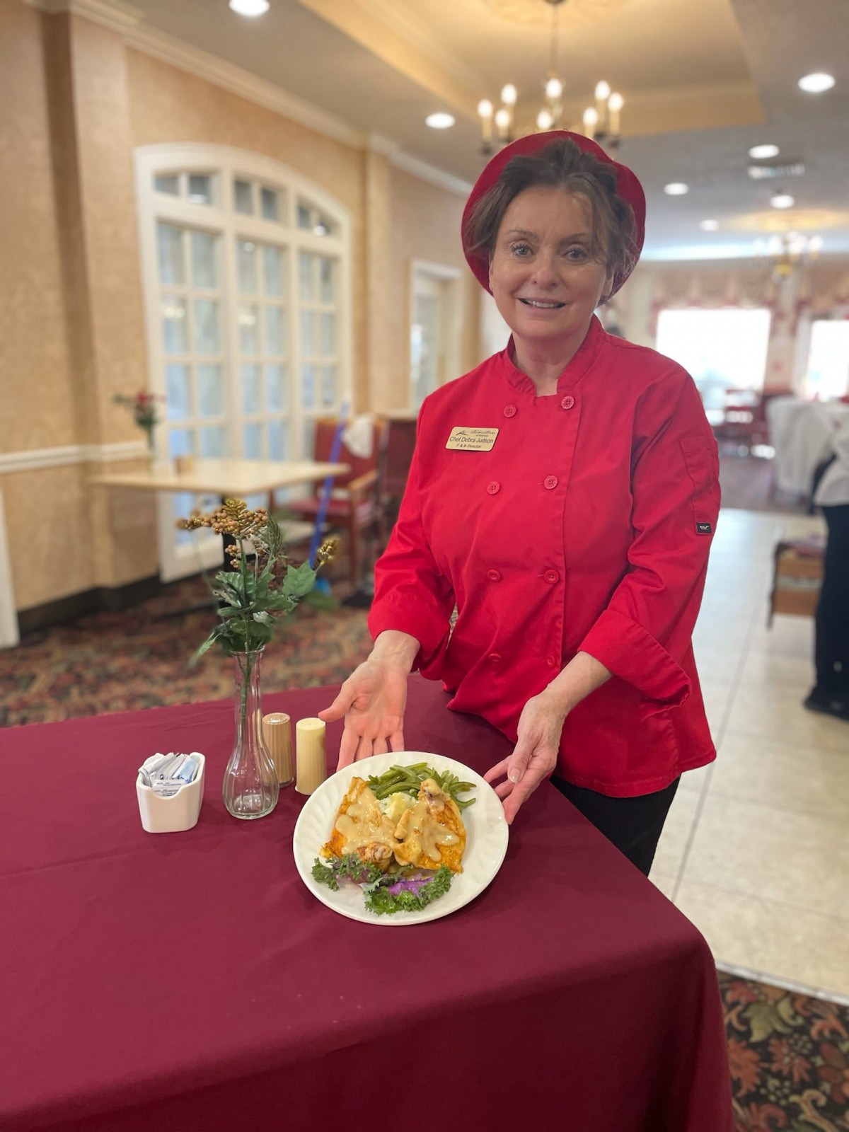 A chef in a red uniform proudly presents a plated dish in a restaurant setting.