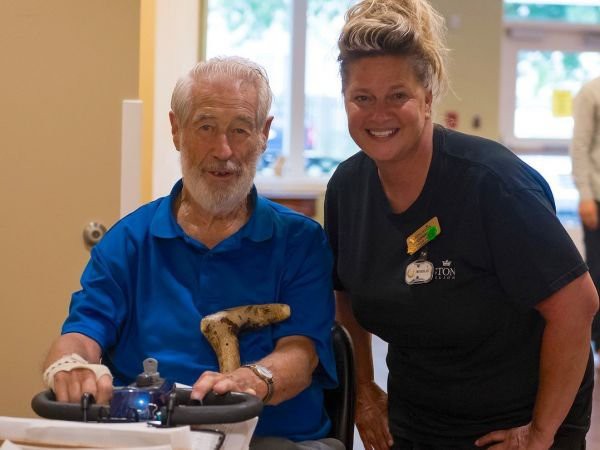 An elderly man in a wheelchair smiling with a caregiver beside him.