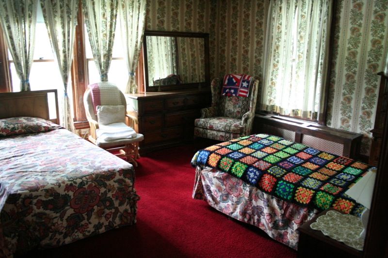 A vintage-style bedroom with red carpeting, featuring a bed, armchair, dresser with mirror, and floral curtains.