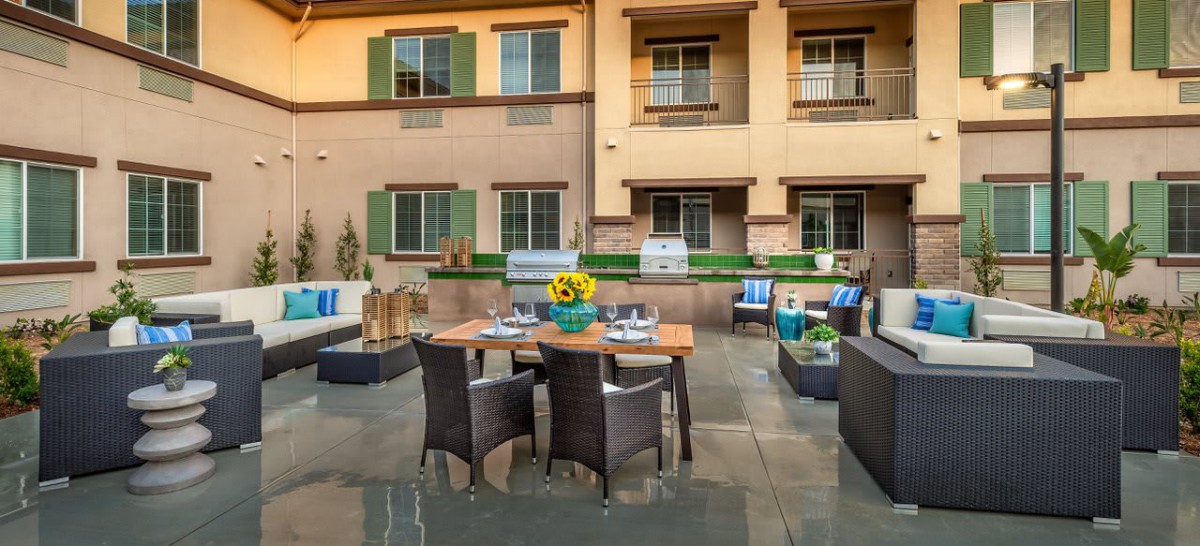A cozy outdoor patio with a dining area featuring wicker chairs, a wooden table adorned with sunflowers, and a building backdrop.