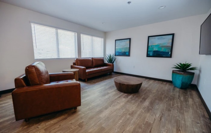 A modern living room featuring brown leather furniture, a round coffee table, and wall art, illuminated by natural light from large windows.
