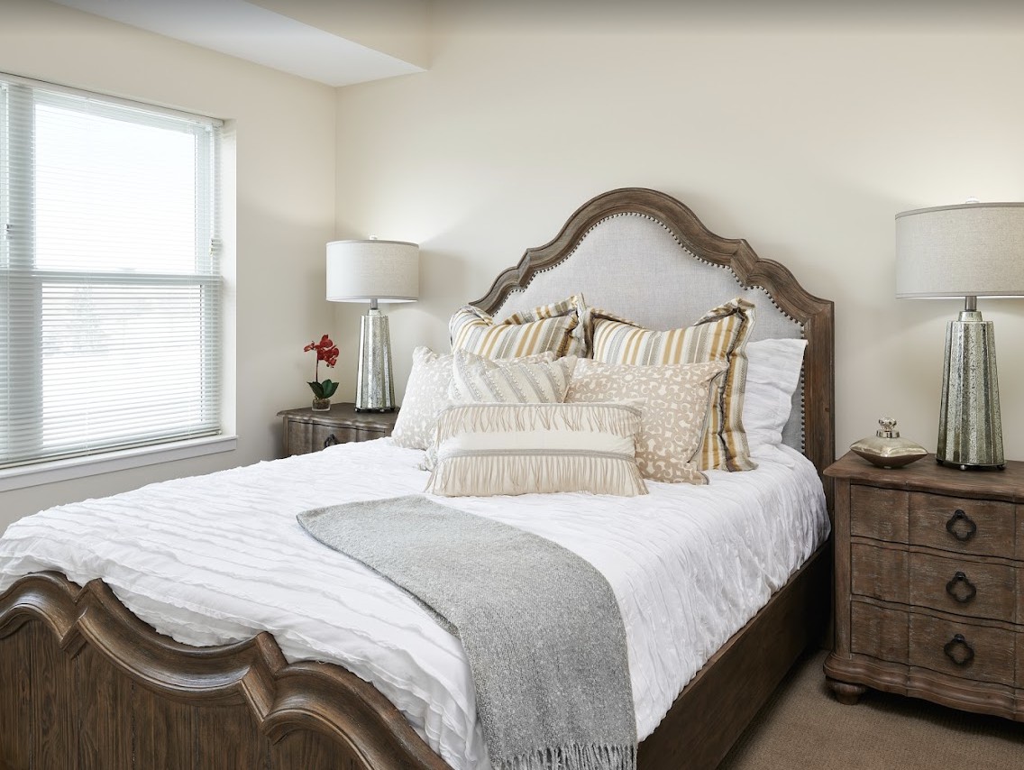 A cozy and elegantly decorated bedroom with a large bed, decorative pillows, a lamp, and a window allowing natural light.