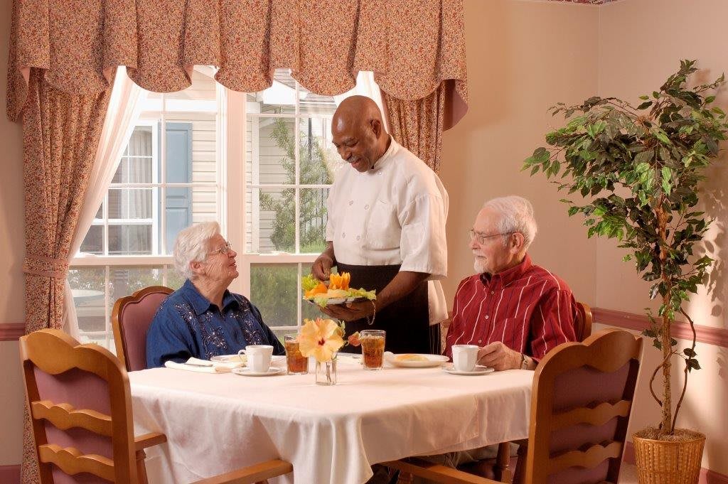 A waiter serves a meal to two seniors at a dining table indoors.