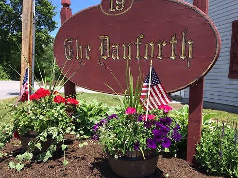 A garden display featuring a sign reading 'The Danforth', surrounded by colorful flowers and a small American flag, on a sunny day.