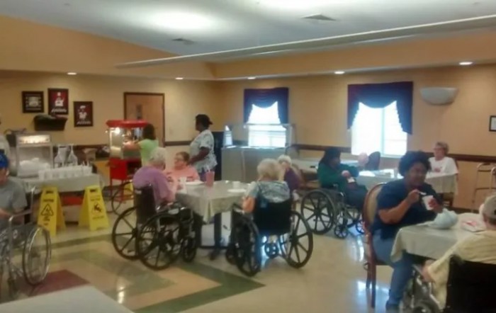 Elderly residents in wheelchairs gathered in a common area of a nursing home with staff members nearby.