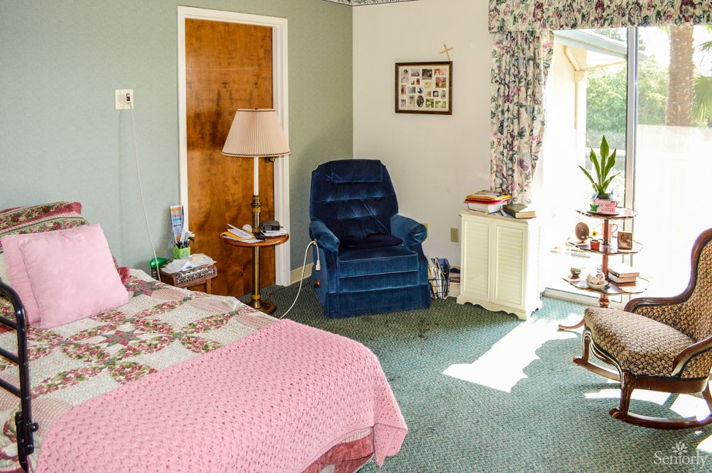 A cozy bedroom with vintage decor, featuring a bed, armchair, lamp, and window with floral curtains.