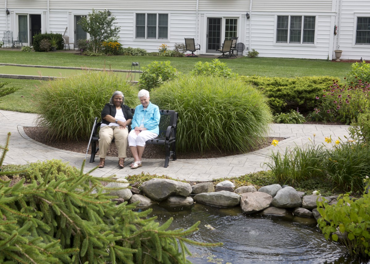 An elderly couple sits on a bench enjoying the serene garden and pond view.