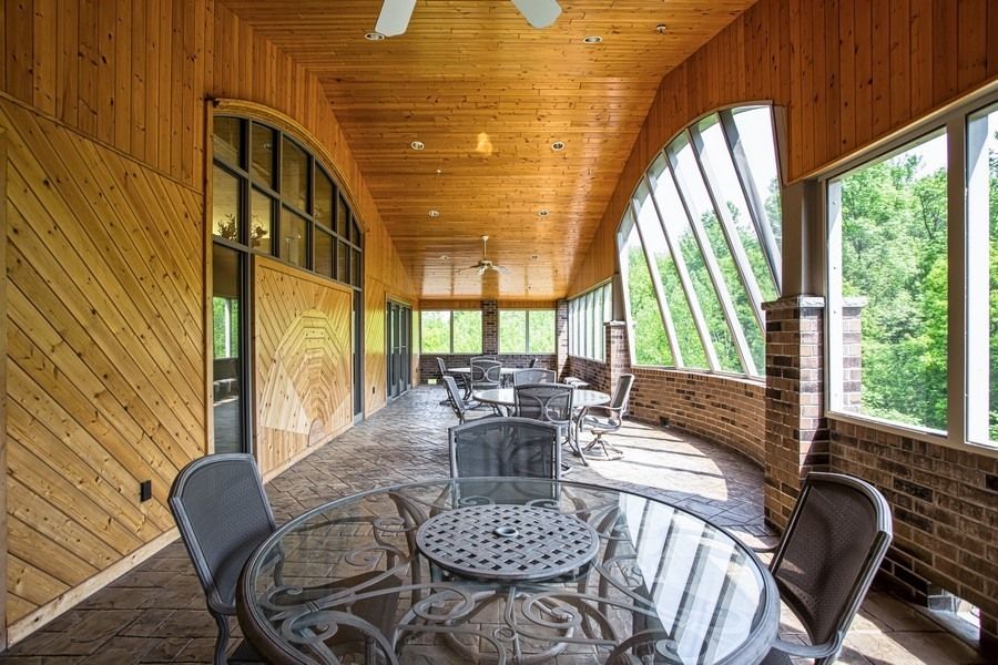 A spacious porch with glass-topped tables and chairs, featuring a wooden ceiling and large windows.