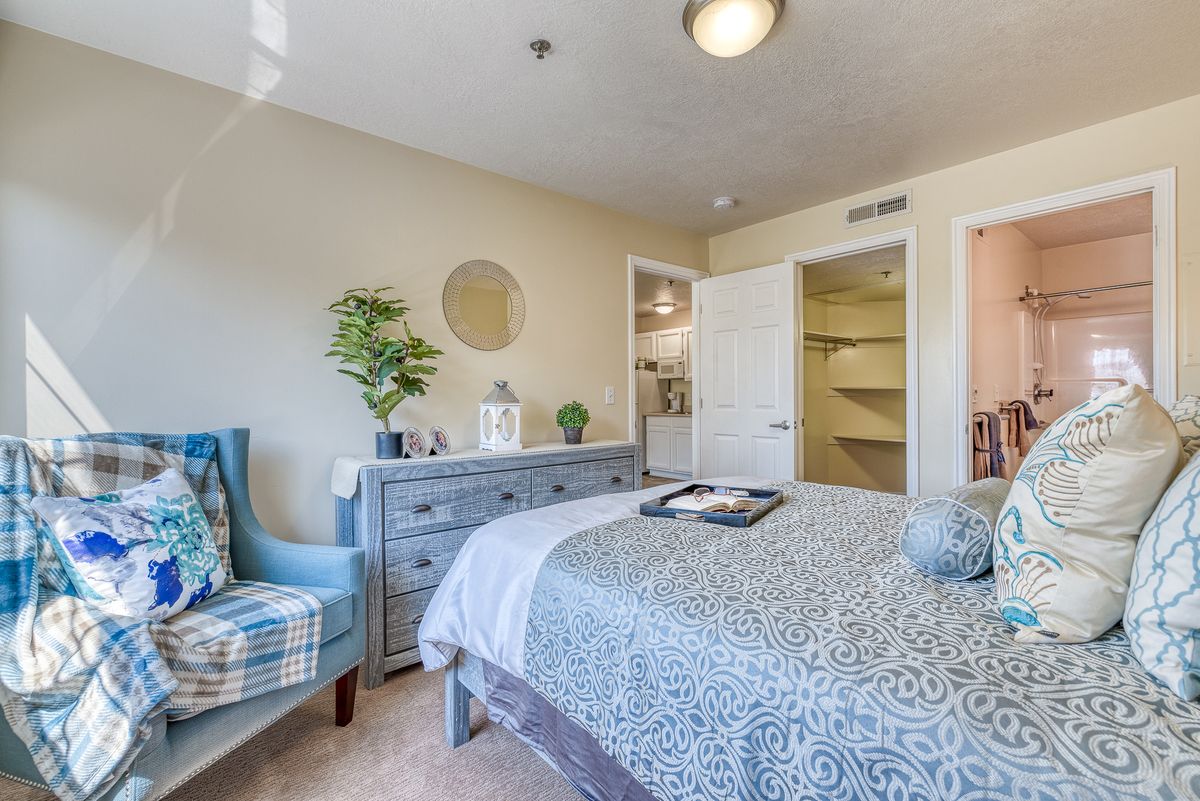 A cozy bedroom with a stylish chair, dresser, and neatly made bed adorned with decorative plants and a round mirror on the wall.