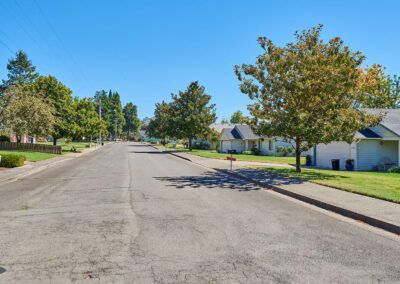 A quiet suburban street lined with trees and houses under a clear blue sky.
