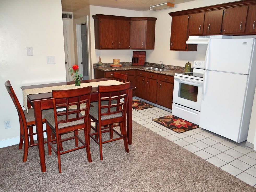 A cozy kitchen with wooden cabinets and a dining area, featuring white appliances and a carpeted floor.