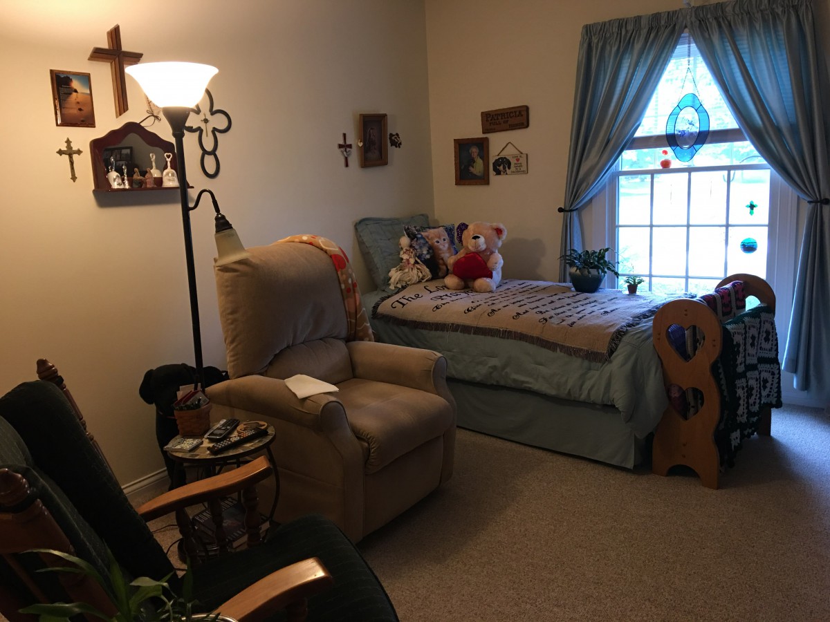 A cozy bedroom with an armchair, a bed adorned with stuffed animals, and decorative items such as crosses and framed pictures on the walls.