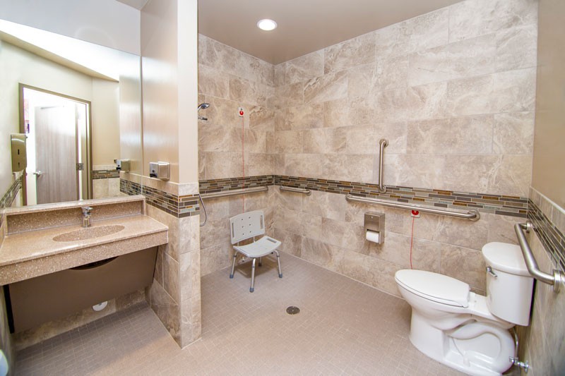 An accessible bathroom featuring a toilet, sink, and safety chair with grab bars installed on the tiled walls.