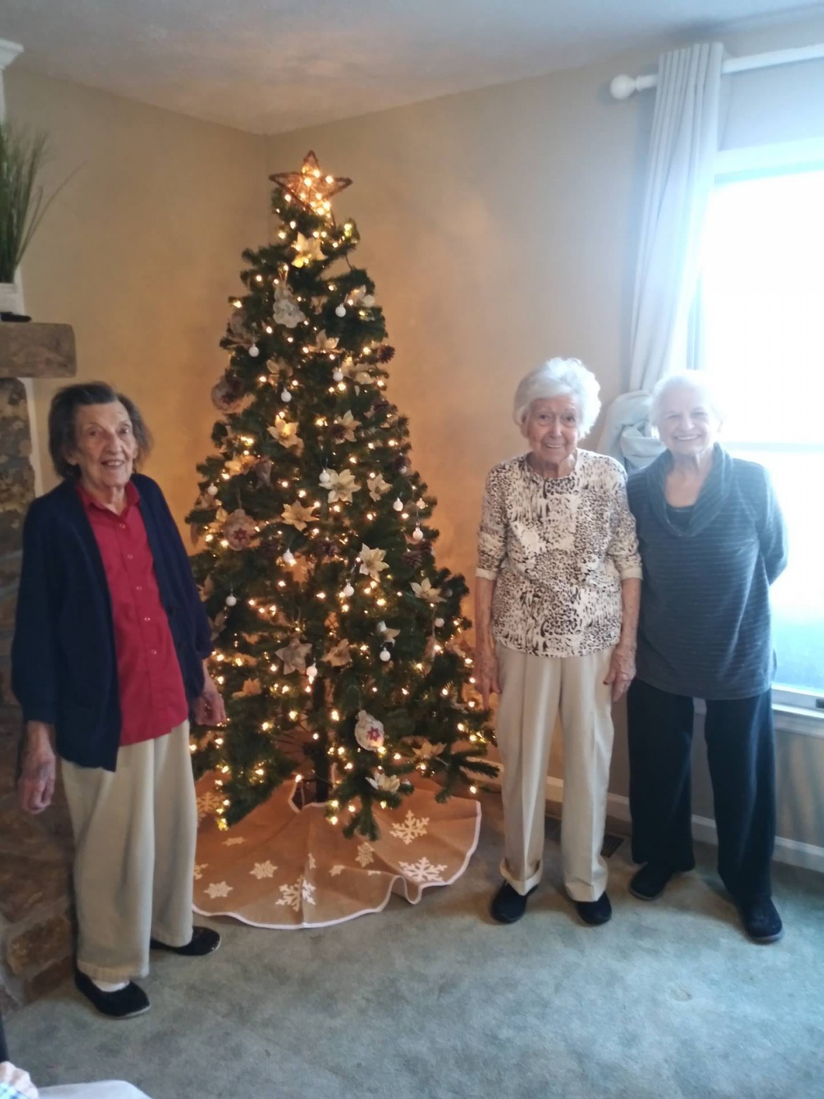 Three elderly women standing by a decorated Christmas tree in a cozy living room.