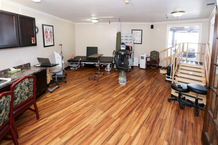 A spacious physical therapy room featuring exercise equipment and treatment tables on a wooden floor.