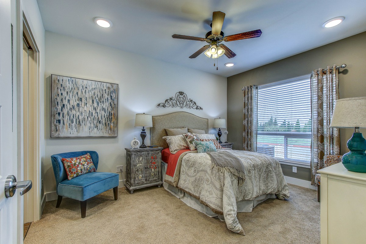A cozy and elegantly furnished bedroom featuring a large bed with decorative pillows, a patterned rug, artwork on the wall, a blue armchair, and a ceiling fan.