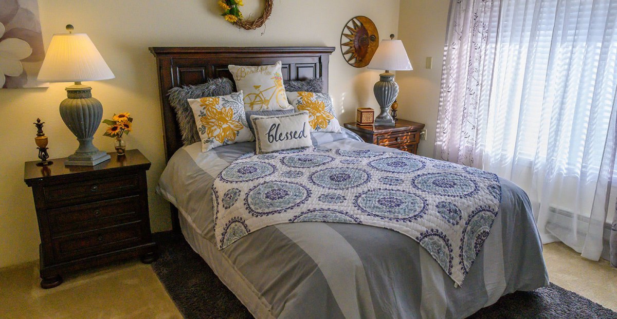 A cozy bedroom with decorative pillows, patterned bedding, and a bedside lamp.