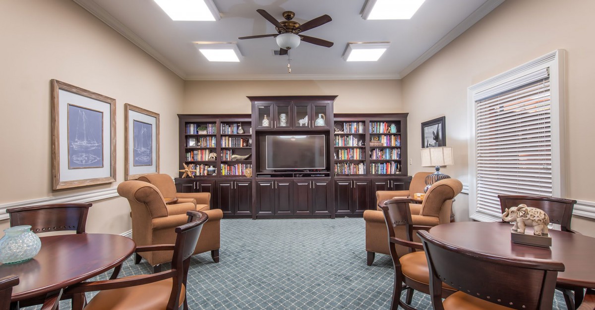A cozy library room with bookshelves, armchairs, a large television, and a ceiling fan.