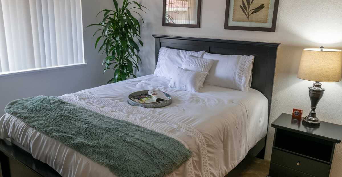 A cozy bedroom with a neatly made bed, featuring white linens, pillows, an indoor plant, and a decorative tray.